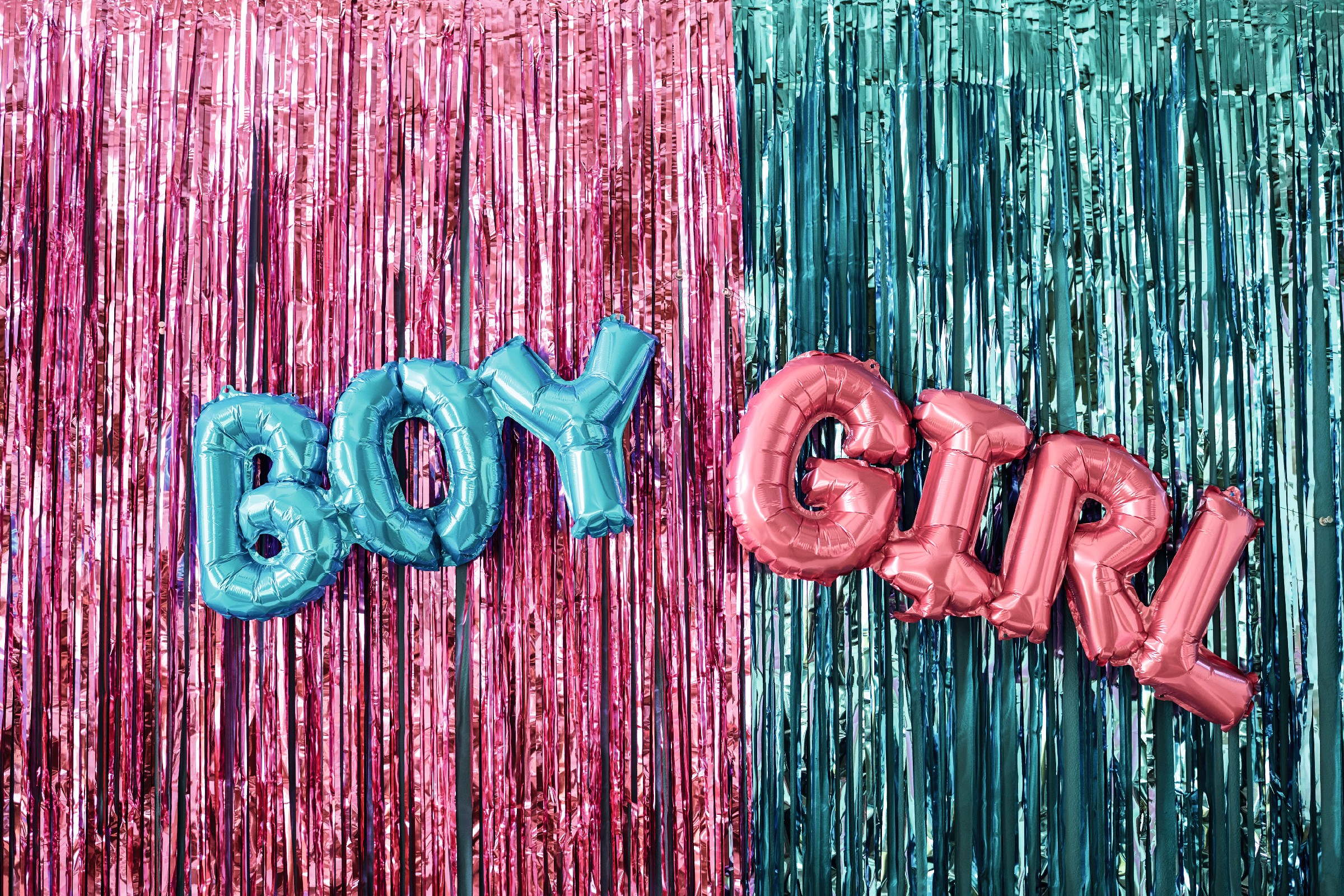 Blue balloon letters spelling boy and pink balloon letters spelling girl hanging against metallic pink and teal tinsel backdrop representing gender reveal party decoration Blue balloon letters spelling boy and pink balloon letters spelling girl hanging against metallic pink and teal tinsel backdrop representing gender reveal party decoration